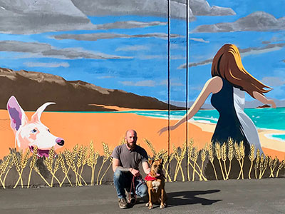 Man with Dog Hunched In Front of Mural of Woman And Dog In Wheat Field
