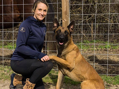 Woman In Navy Top Holding The Paw Of A Large Dog While Crouched In Front Of A Fence