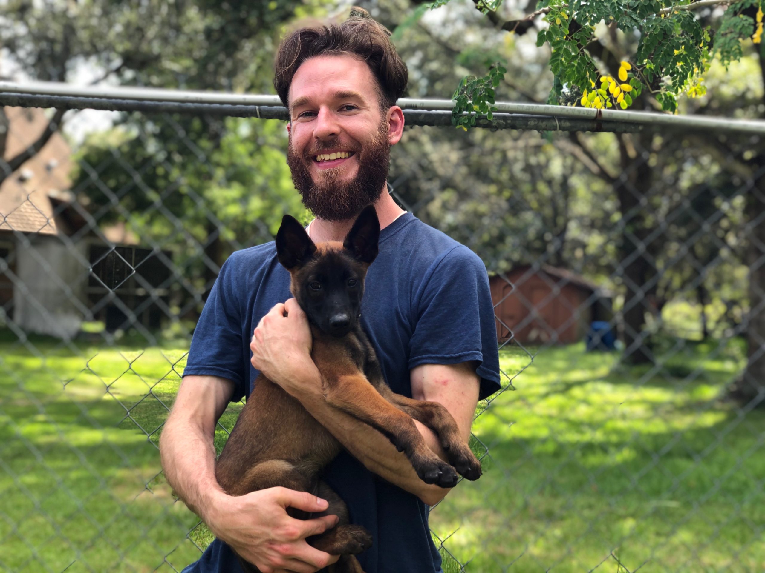 Man With Beard Standing Against Fence Holding Puppy In His Arms