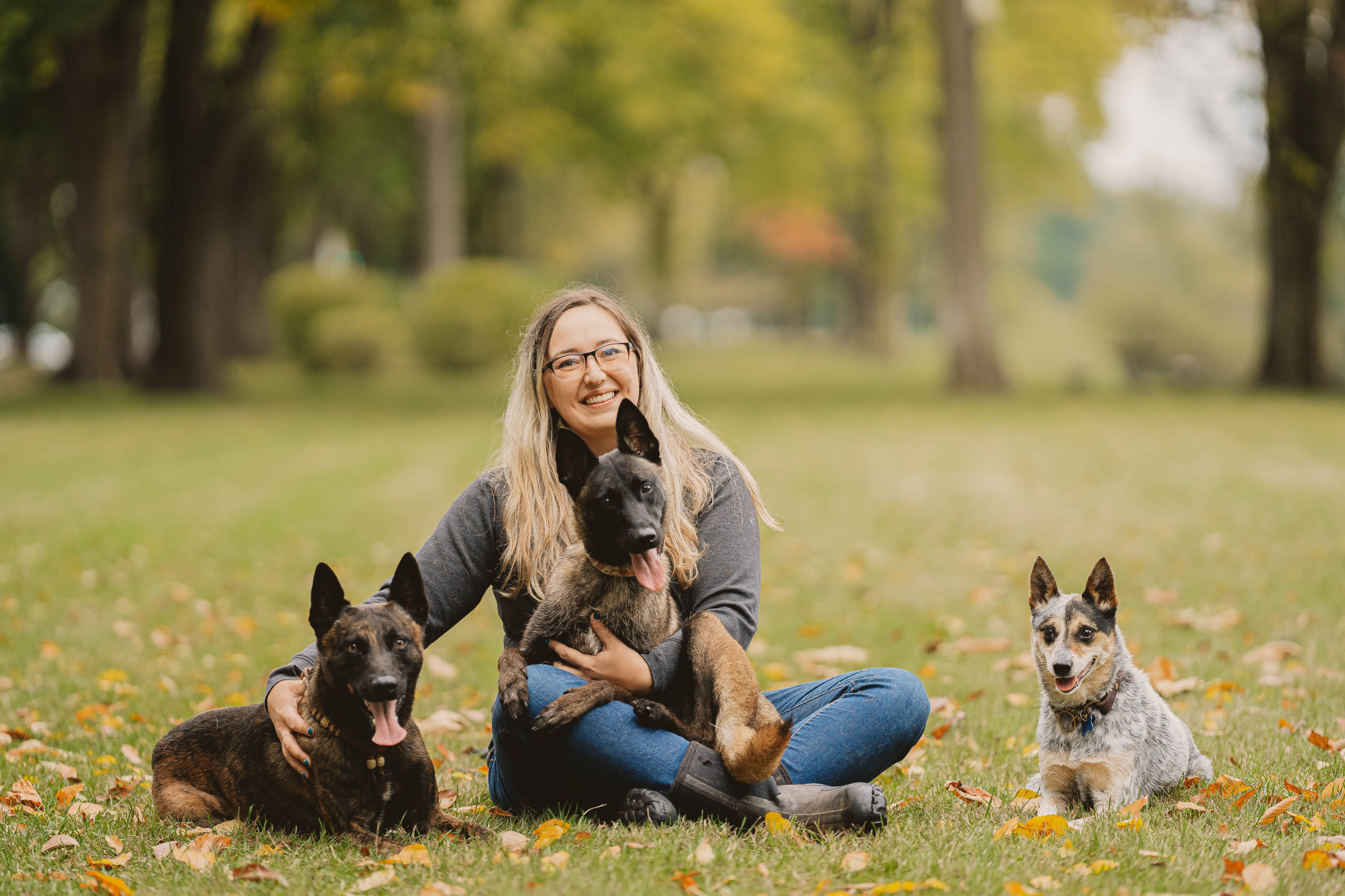 Blond Woman With Glasses Sitting In Park Grass With One Dog On Her Lap And Two Others Beside Her