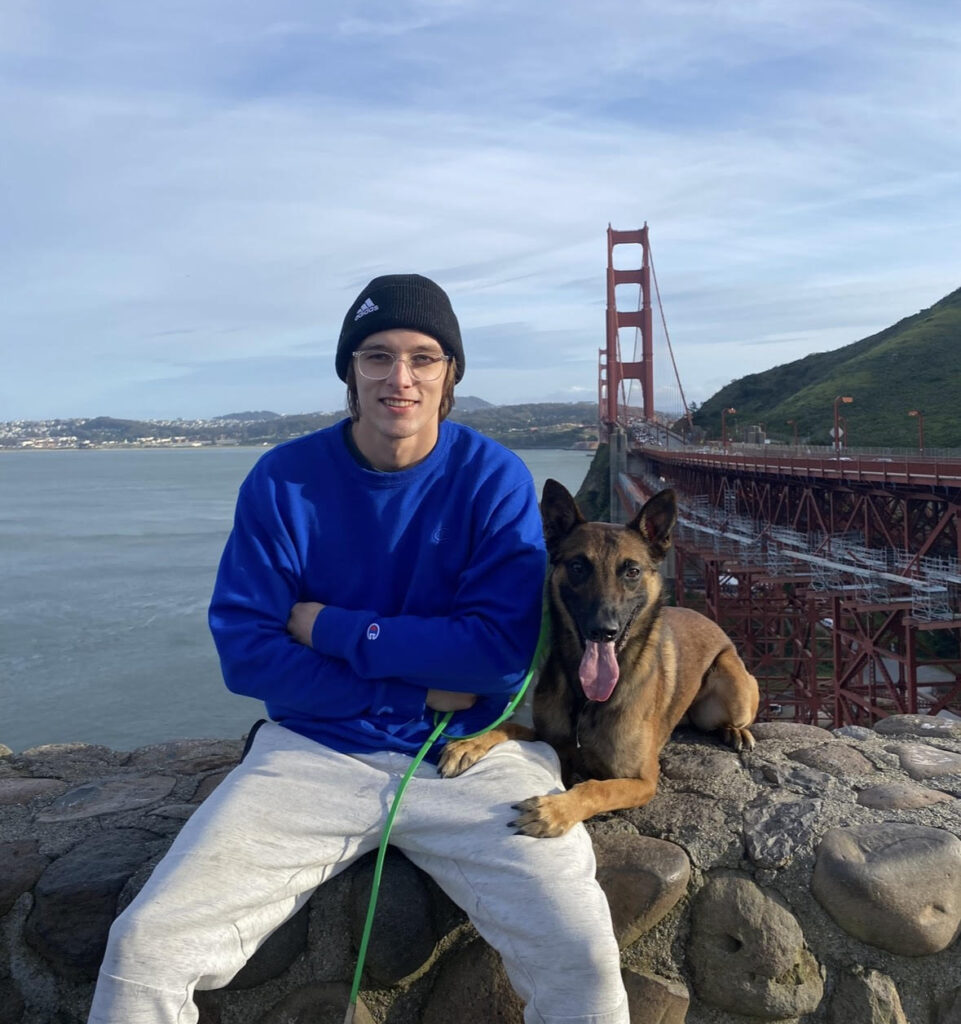 Man With Blue Sweatshirt Sitting On Stone Ledge Next To Golden Gate Bridge With Dog