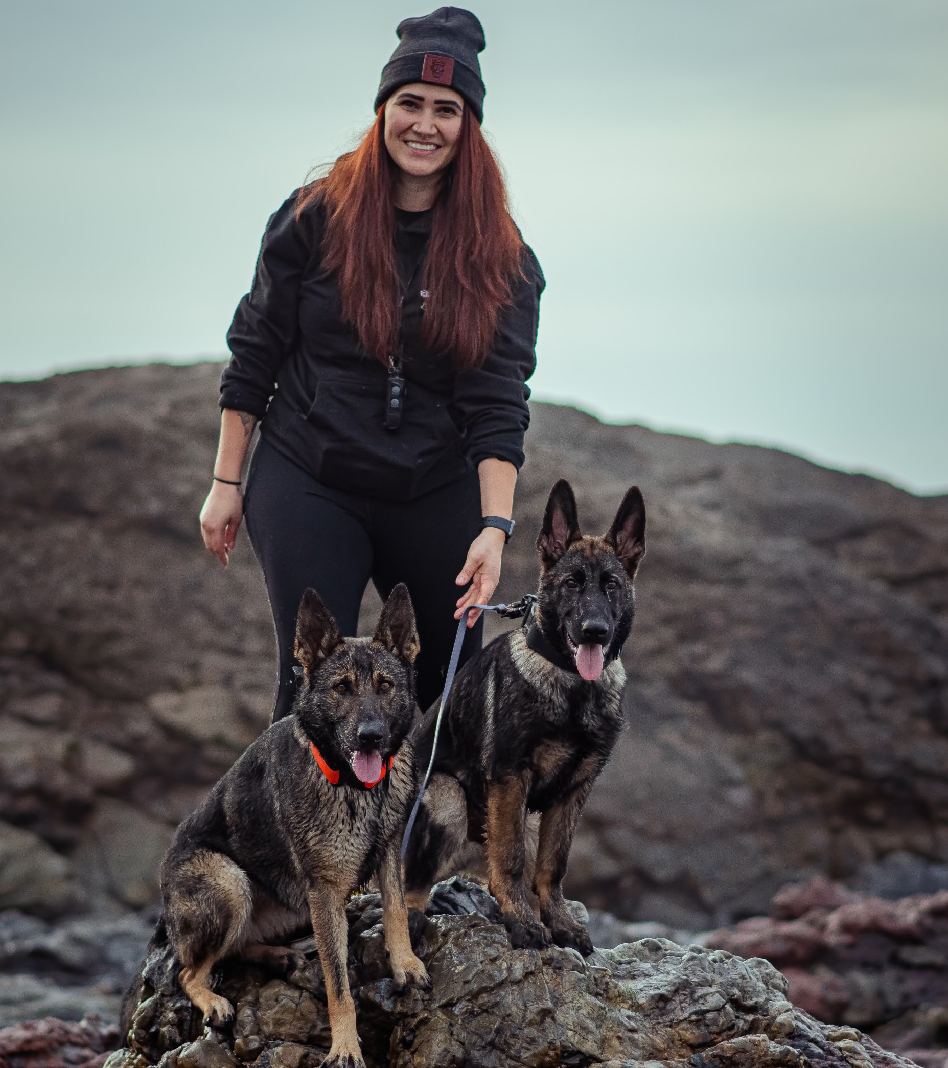 Red-Headed Woman Wearing A Beanie Standing On A Black Rock With Two Large Dogs