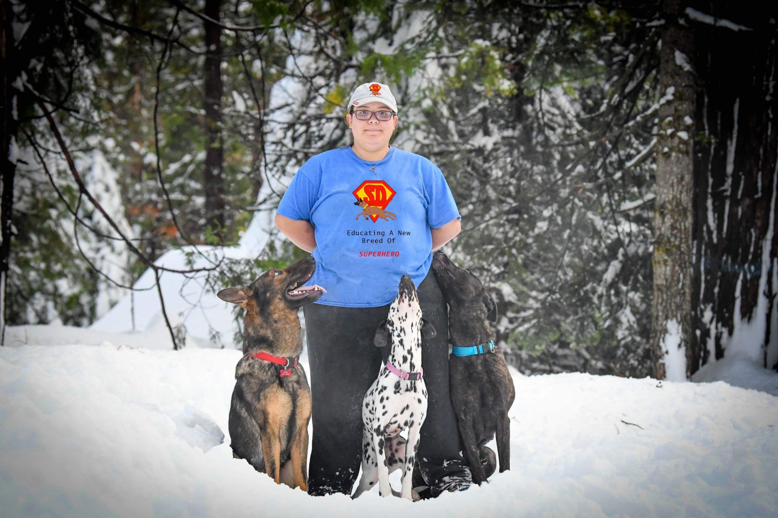 Woman With Glasses And Blue T-Shirt Standing In Snow With Three Dogs Looking Up At Her