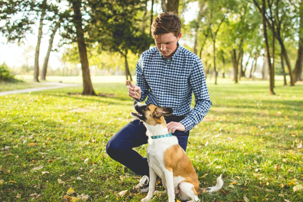 Man In Checked Shirt Kneeling Next To His Seated Dog And Holding On To His Collar