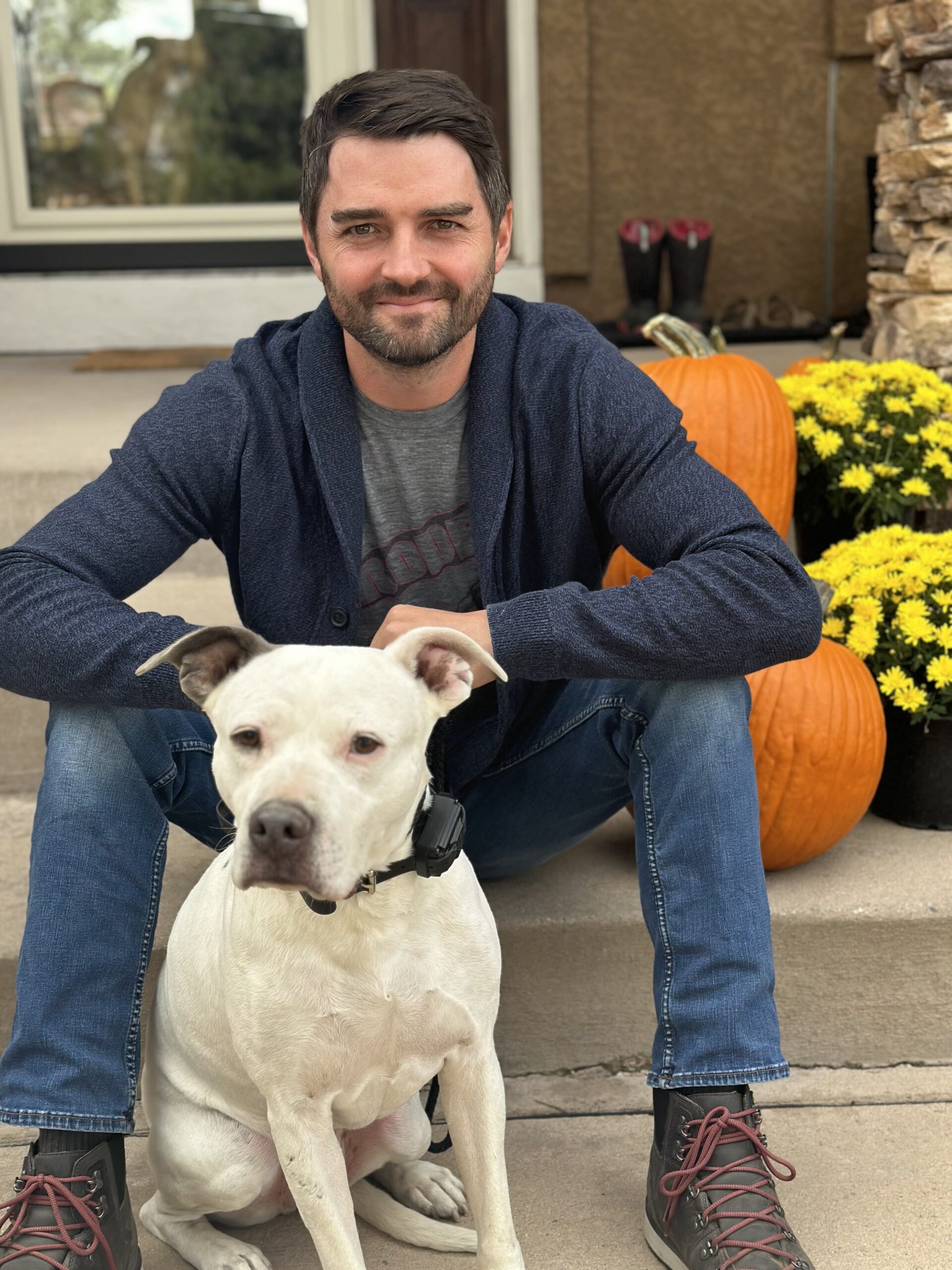 Man Sitting On Front Steps Of Home With White Pitbull And Two Orange Pumpkins