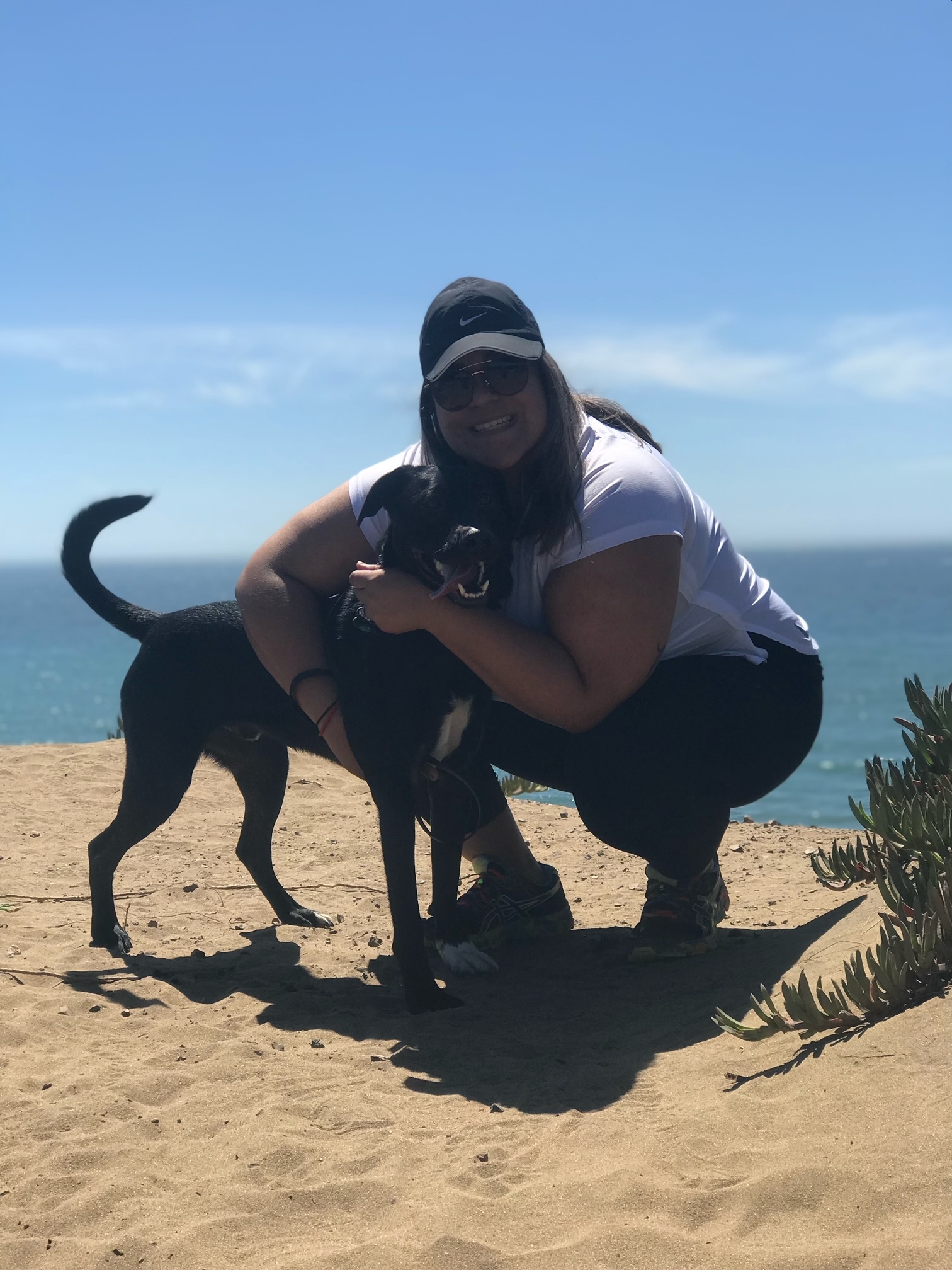 Woman With Black Cap Crouched And Hugging Her Black Dog At The Beach